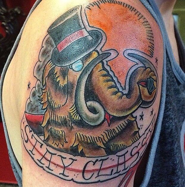 Unusual colorful mammoth in hat with ribboned lettering tattoo on shoulder
