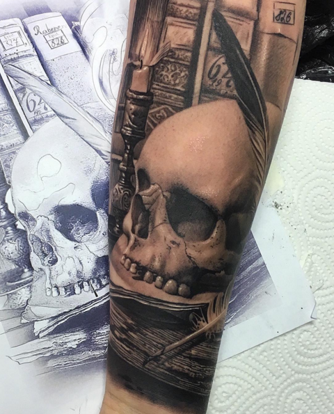 Black and gray style detailed arm tattoo of human skull with feather and books