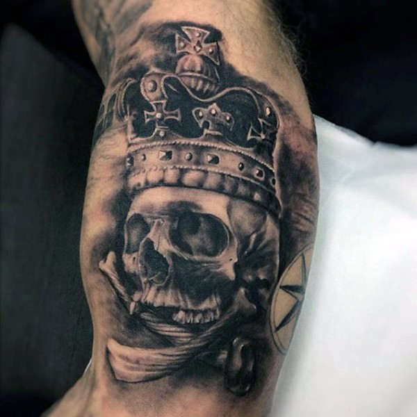 Black and gray style skull with crown tattoo on biceps combined with crossed bones