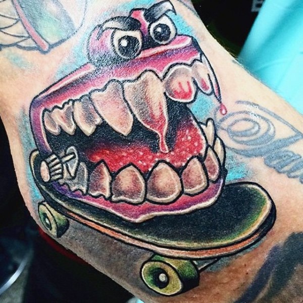 Cartoon style colored evil jaw with skateboard tattoo on arm