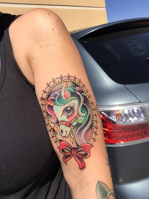 Cute little cartoon unicorn portrait tattoo on arm stylized with red bow