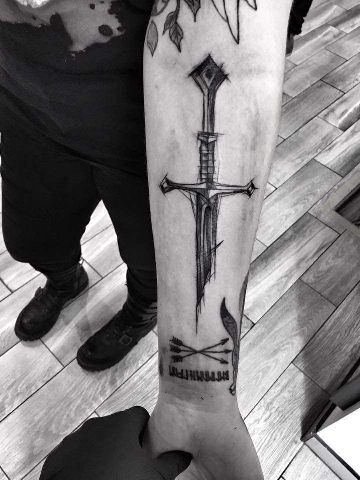 Tinta preta estilo fantasia pintada por Inez Janiak antebraço tatuagem de espada quebrada com letras
