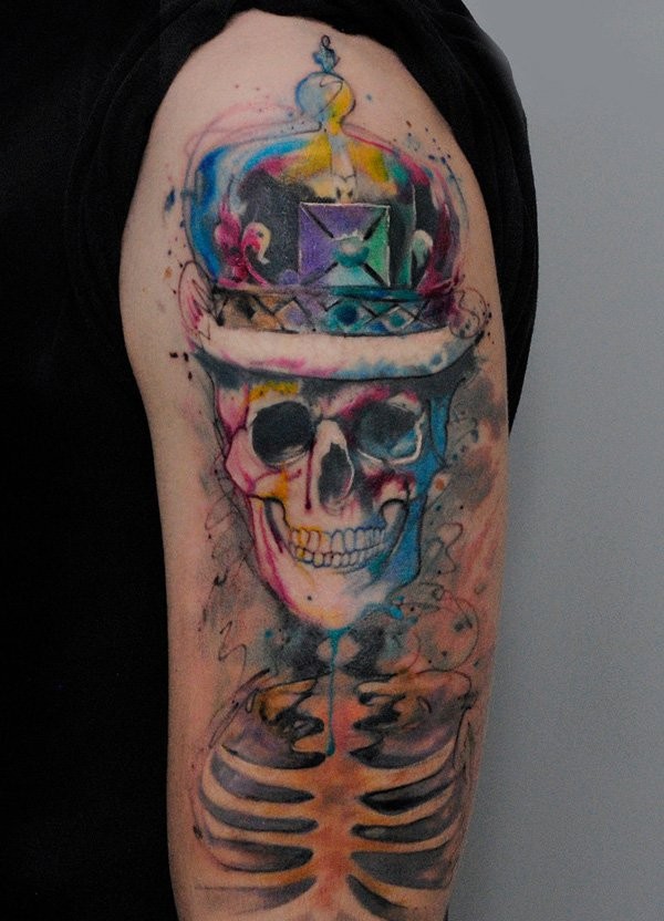 Funny smiling skeleton with monarchy crown colored tattoo on shoulder with watercolor elements
