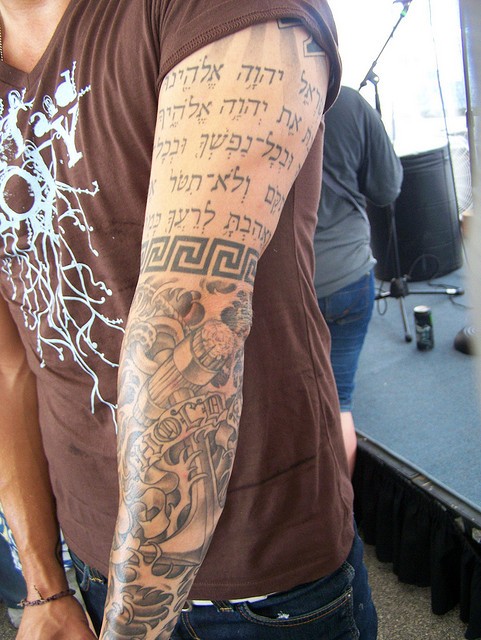 Hebräischer Schriftzug, schwarzes Armband und riesiger abgeseilter Anker in Wasserwellen blasses Ärmel Tattoo