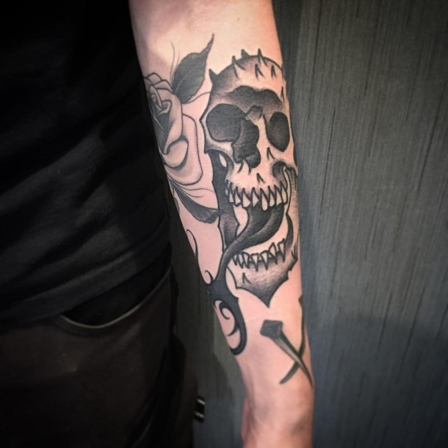 Illustrative style black ink forearm tattoo of human skull with flower