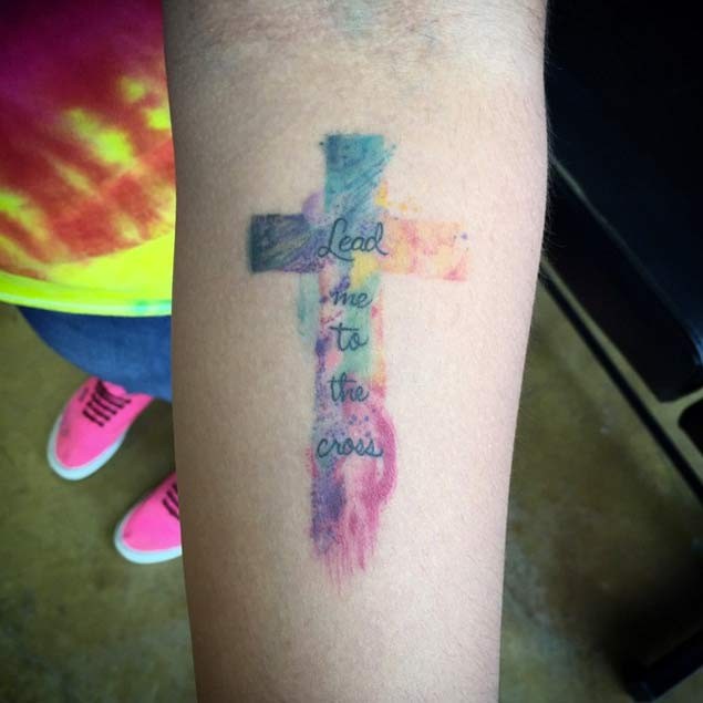 Tatuaje en el antebrazo, cruz multicolor con inscripción diminuta