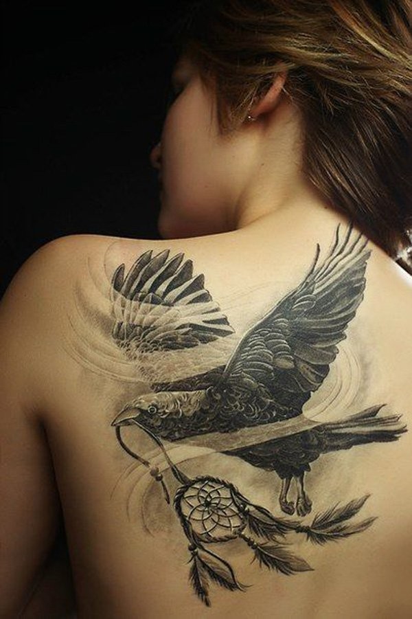 Tatuaje en el omóplato, cuervo volando  con atrapasueños en el pico