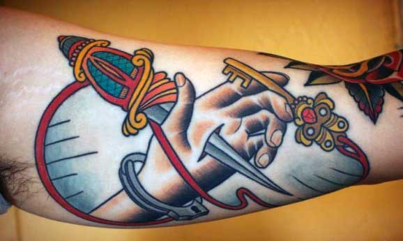 Tatuaje en el brazo, mano perforada por daga y llave, estilo old school de varios colores