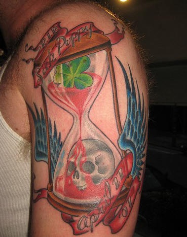 el tatuaje conmemorativo de una calavera y la sangre dentro de un reloj de arena con alas hecho en color en el brazo