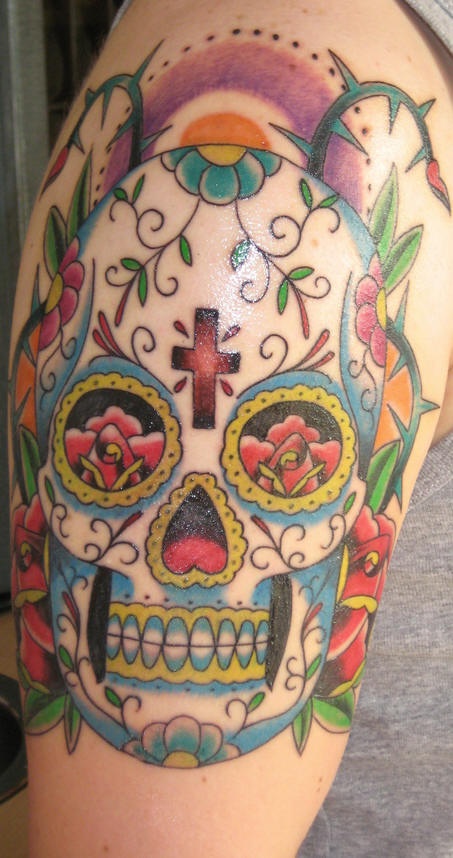 el tatuaje de una calavera mexicana con una cruz en sy frente hecho en el hombro con tinta de varios colores