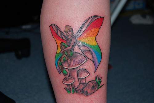 Tatuaje de una hada con alas color arco iris sentada sobre setas