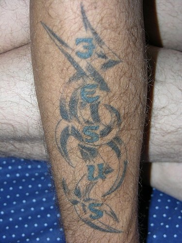 Tatuaje en el antebrazo, inscripción Jesús entre ornamento curvado