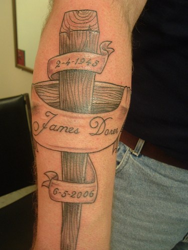 Tatuaje en el antebrazo, estaca de madera, cinta con nombre y fecha
