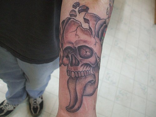 Tatuaje en el antebrazo, cráneo con lengua larga y con cabeza rota