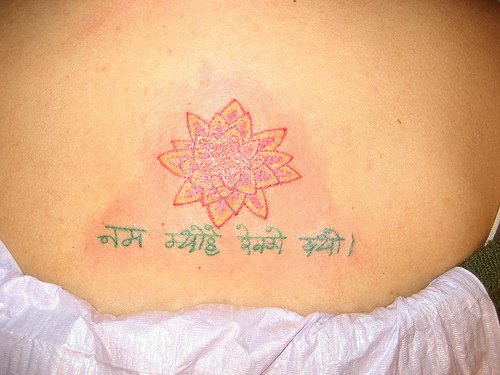 el tatuaje chico de una flor de loto de color naranja con una escritura en hindu