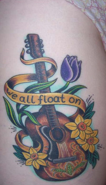 el tatuaje muy bonito de una guitarra con flores y un liston con una frase hecho en color