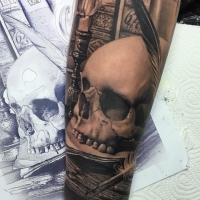 Black and gray style detailed arm tattoo of human skull with feather and books