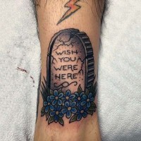Cartoonischer kleiner farbiger Grabstein mit Schriftzug und Blumen Tattoo am Knöchel