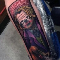Funny cartoon style colored Joker with lettering tattoo on forearm