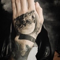 Gray washed style detailed hand tattoo of small skull with lettering