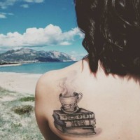 Steaming cup on pale of books detailed black and white upper back tattoo