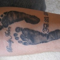 Tattoo von schwarzen männlichen Fußspuren mit Namen und Datum am Unterarm
