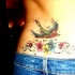 Le tatouage sur bas du dos avec un moineau multicolore en vol au dessous de fleurs