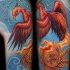 el tatuaje de un ave Fenix detallado con una traceria aprededor hecho en el fondo azul en el hombro