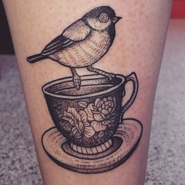 Tiny black ink natural looking bird sitting on cup tattoo on ankle stylized with flowers