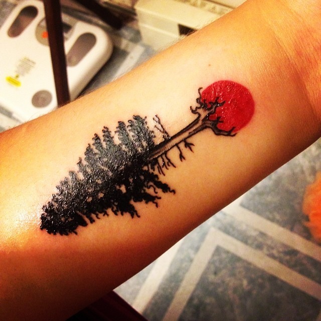 Tiny black ink tree tattoo on forearm with red sun